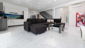 Ein modernes Wohnzimmer mit einem Ecksofa, einem Glastisch und einer Küchenzeile mit weißen Schränken und einer Mikrowelle.
