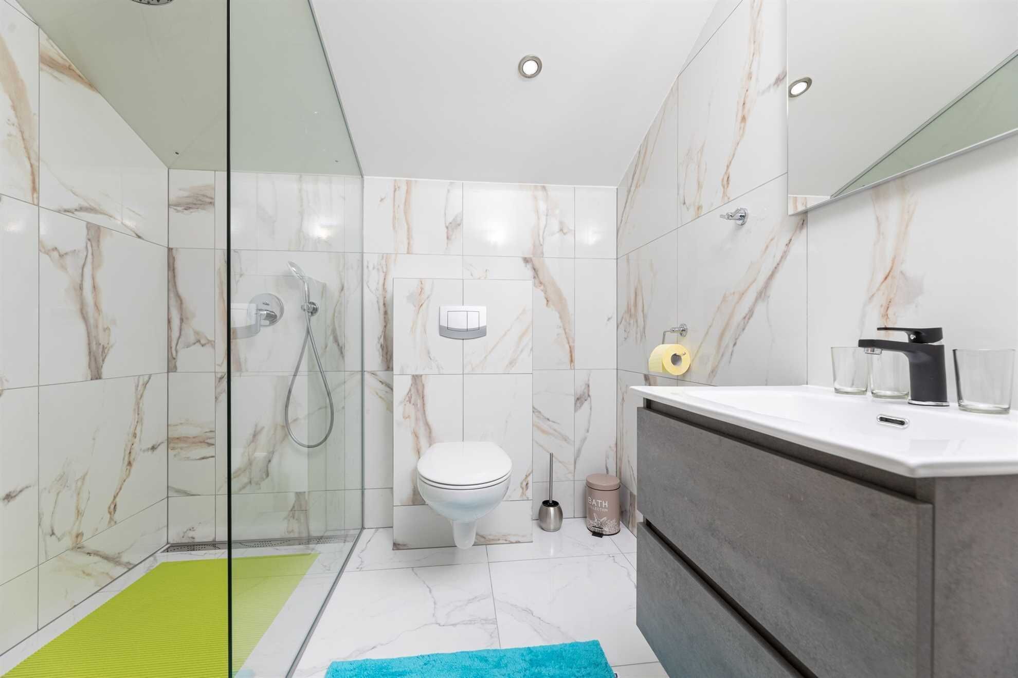 Een moderne badkamer met een douchecabine van glas, een toilet, een wastafel met spiegel en een groene badmat.