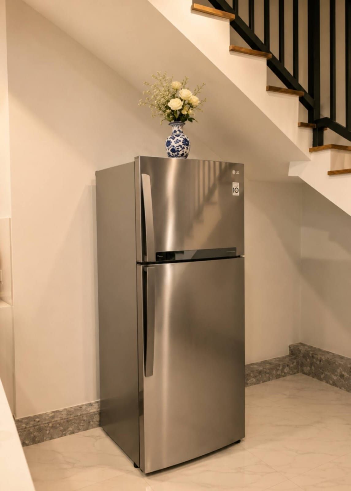 A stainless steel refrigerator with a vase of flowers on top, located in a corner of a room with a tiled floor and a staircase in the background.