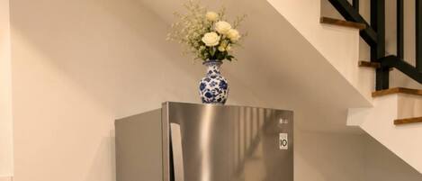 A stainless steel refrigerator with a vase of flowers on top, located in a corner of a room with a tiled floor and a staircase in the background.