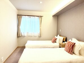 Quarto de hotel com duas camas, uma janela com cortinas, uma mesa de cabeceira e uma luminária fixada na parede.