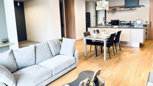 A modern living room with a grey sofa, a dining table with chairs, a kitchen area with a stove and sink, and a wooden floor.