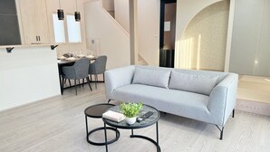 Un salon moderne avec un canapé gris, deux petites tables rondes et un coin repas à l’arrière-plan.