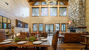 A spacious dining area with wooden tables and chairs, large windows, and a stone fireplace. - Swimming Beaver Mansion · Indoor Pool Year-Round, Sleeps 28, Game Room, Views! (Broken Bow)