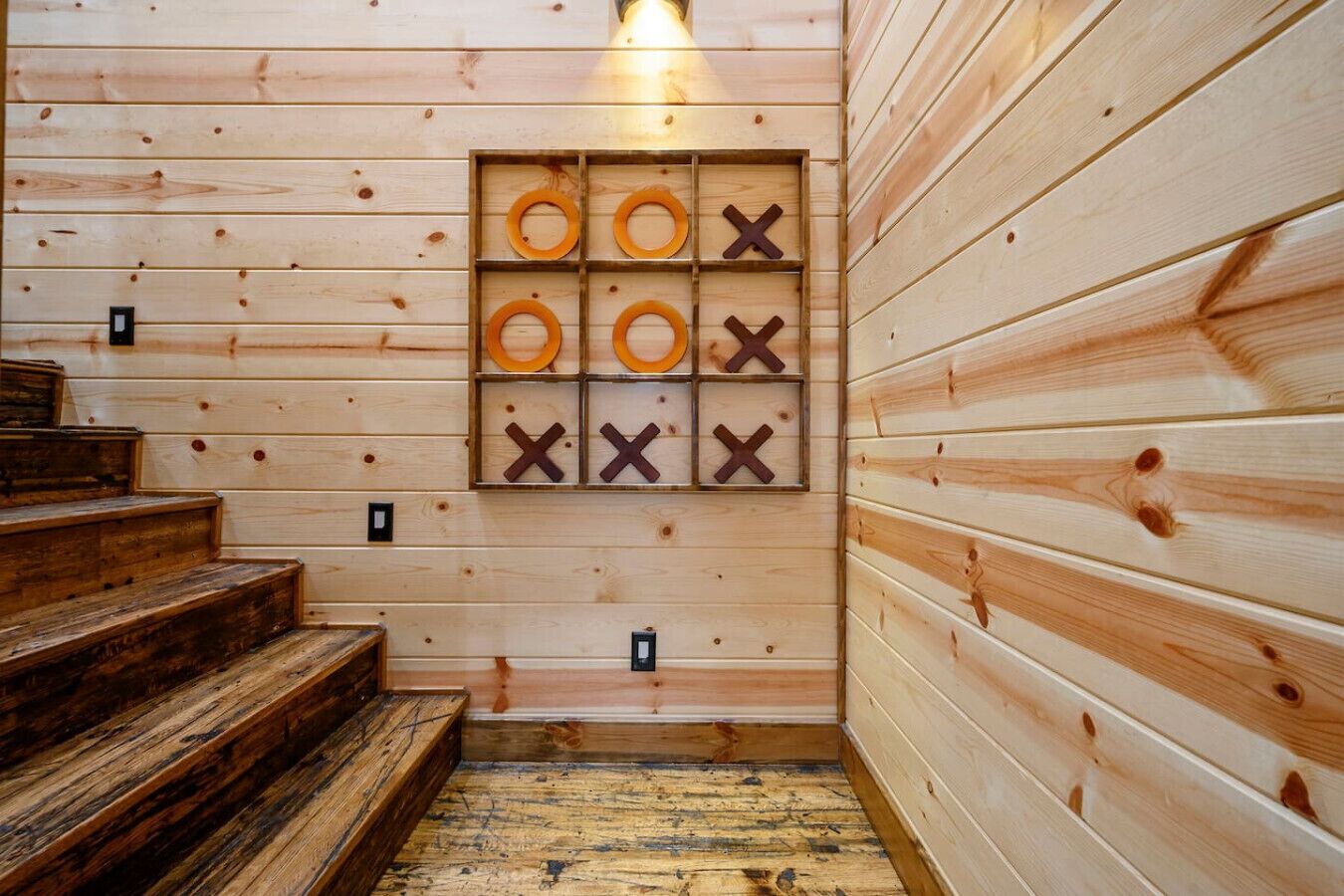 Interior de una cabaña de madera con una escalera de madera, un tablero de juego en la pared y una lámpara.