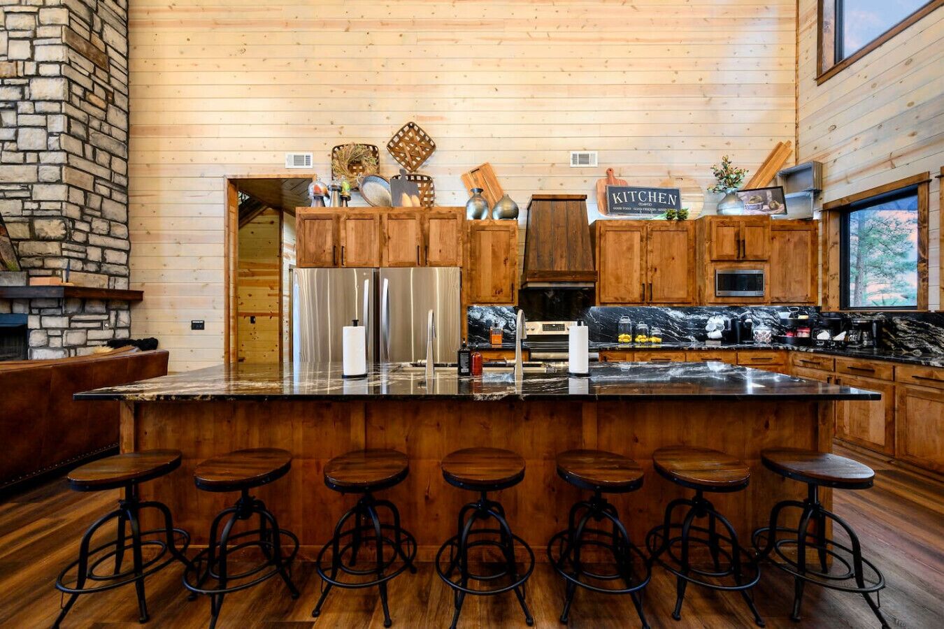 A kitchen with wooden cabinets, a large island with stools, stainless steel appliances, and a stone fireplace.