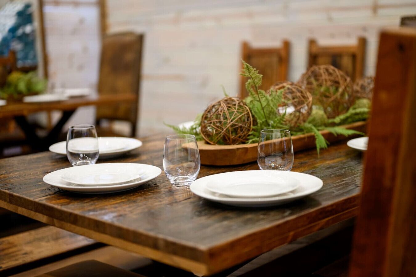Una mesa de comedor de madera con platos blancos y vasos transparentes, rodeada de decoración rústica que incluye esferas tejidas y follaje.