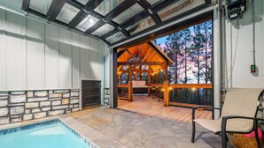 A pool area with a stone deck, a wooden deck with a covered seating area, and a view of a forested landscape. - Swimming Beaver Mansion · Indoor Pool Year-Round, Sleeps 28, Game Room, Views! (Broken Bow)