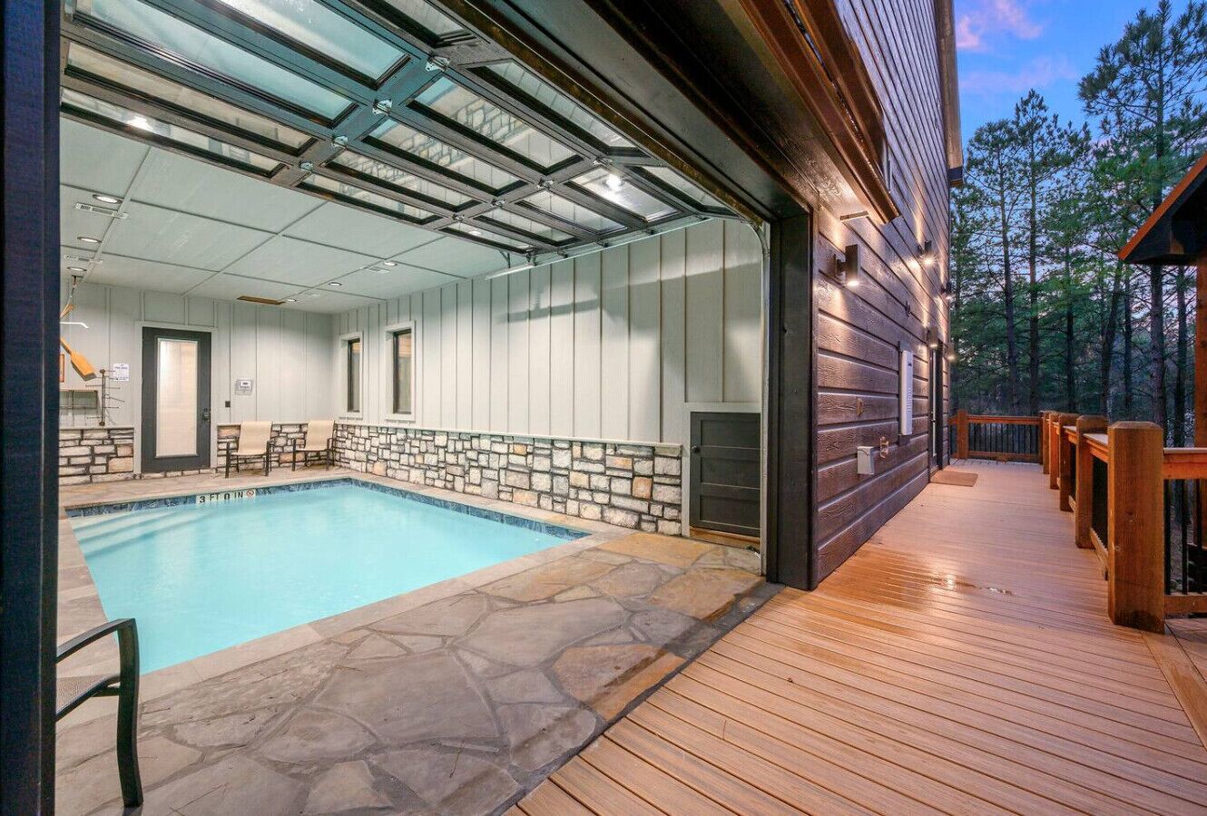 Una piscina con baldosas de piedra, una terraza de madera y una sala acristalada con pared de piedra.