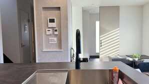 Een moderne hotelkamer met een geïntegreerde keukenbar, een bedieningspaneel aan de muur en een zithoek met een tafeltje en een plant.