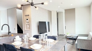 Een moderne eetruimte met een rechthoekige tafel voor vier personen, een zwarte stoel, een grijze bank en een televisie aan de muur.