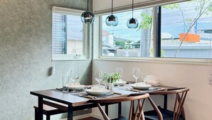 Uma área de refeições com uma mesa posta para quatro pessoas, três luminárias pendentes e uma grande janela com vista para edifícios.