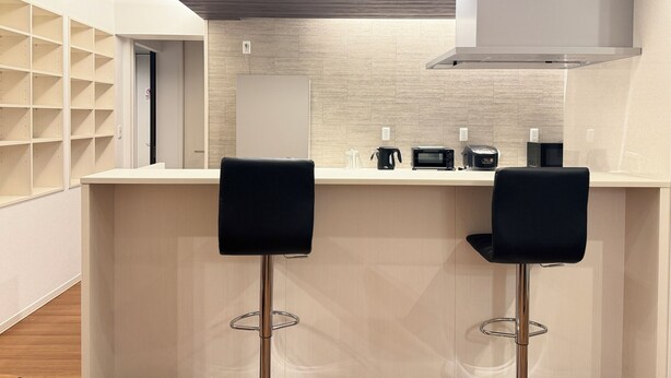 Uma cozinha moderna com bancada branca, dois bancos altos pretos, um exaustor de aço inoxidável e um painel de azulejos.