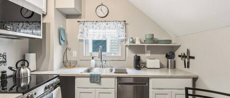 A compact kitchen with a stove, sink, and dishwasher, a clock on the wall, and a window with a patterned curtain.