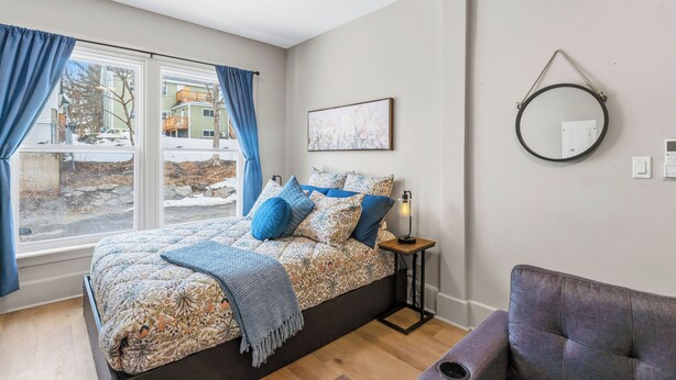 Une chambre à coucher comprenant un lit, une table de chevet, une chaise, une fenêtre avec des rideaux, un miroir fixé au mur et un tableau encadré.