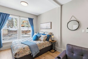 A bedroom with a bed, a nightstand, a chair, a window with curtains, a wall-mounted mirror, and a framed picture. - Gateway to Annapolis Valley | Windsor Studio Suite (Windsor)