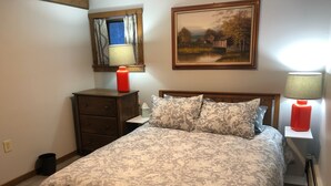 Een slaapkamer met een bed, nachtkastje, kapstok, raam met gordijnen, schilderij aan de muur en een lamp.