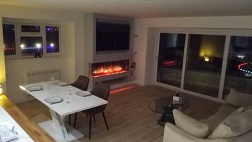 A modern living room with a fireplace, a dining area with a white table, and a large window with a view of the city at night.