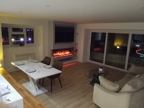 A modern living room with a fireplace, a dining area with a white table, and a large window with a view of the city at night.