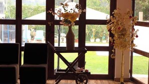 A room with large windows, a cart with a vase of flowers, and a tall vase with flowers.