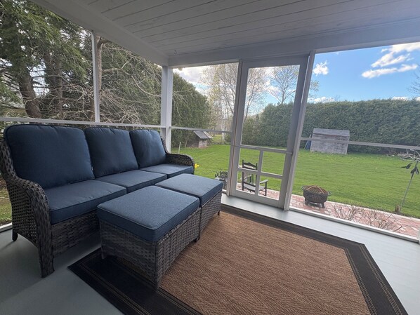 Une terrasse couverte avec un canapé en osier, un tapis marron et une vue sur une pelouse verdoyante et des arbres.