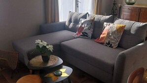 Ett vardagsrum med en grå soffa, ett litet runt bord med en växt, en flätad stol och ett träskåp.