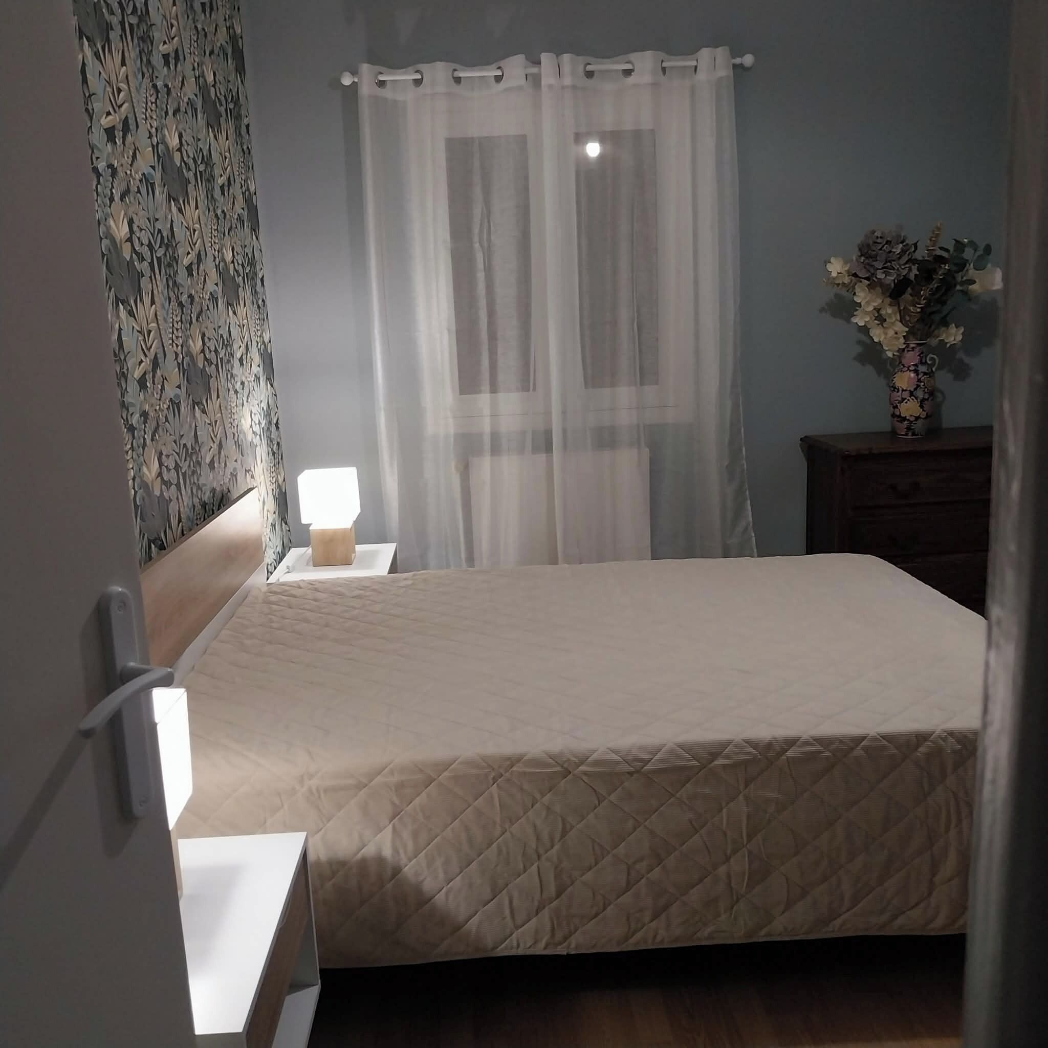 Quarto com cama, mesa de cabeceira com abajur, criado-mudo e parede com padrão floral.