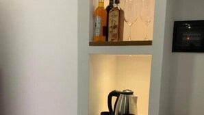 A kitchen area with a coffee maker, two black mugs, a small refrigerator, and a shelf with bottles and glasses. - Vladimir Estate (Gjirokastër)