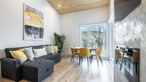 Una sala de estar moderna con un sofá gris oscuro, cojines amarillos, una mesa de comedor de madera con sillas amarillas y un ventanal con vistas a árboles.