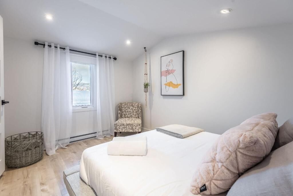 A bedroom with a bed, a chair, a window with curtains, a framed picture on the wall, and a basket on the floor.