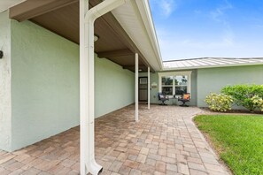 Ein überdachter Patio-Bereich mit Backsteinböden, einer Sitzecke mit Stühlen und Tisch sowie ein kleiner Garten mit Sträuchern.