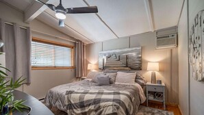 A bedroom with a bed, a ceiling fan, a window with blinds, a painting on the wall, and a bedside table with a lamp.