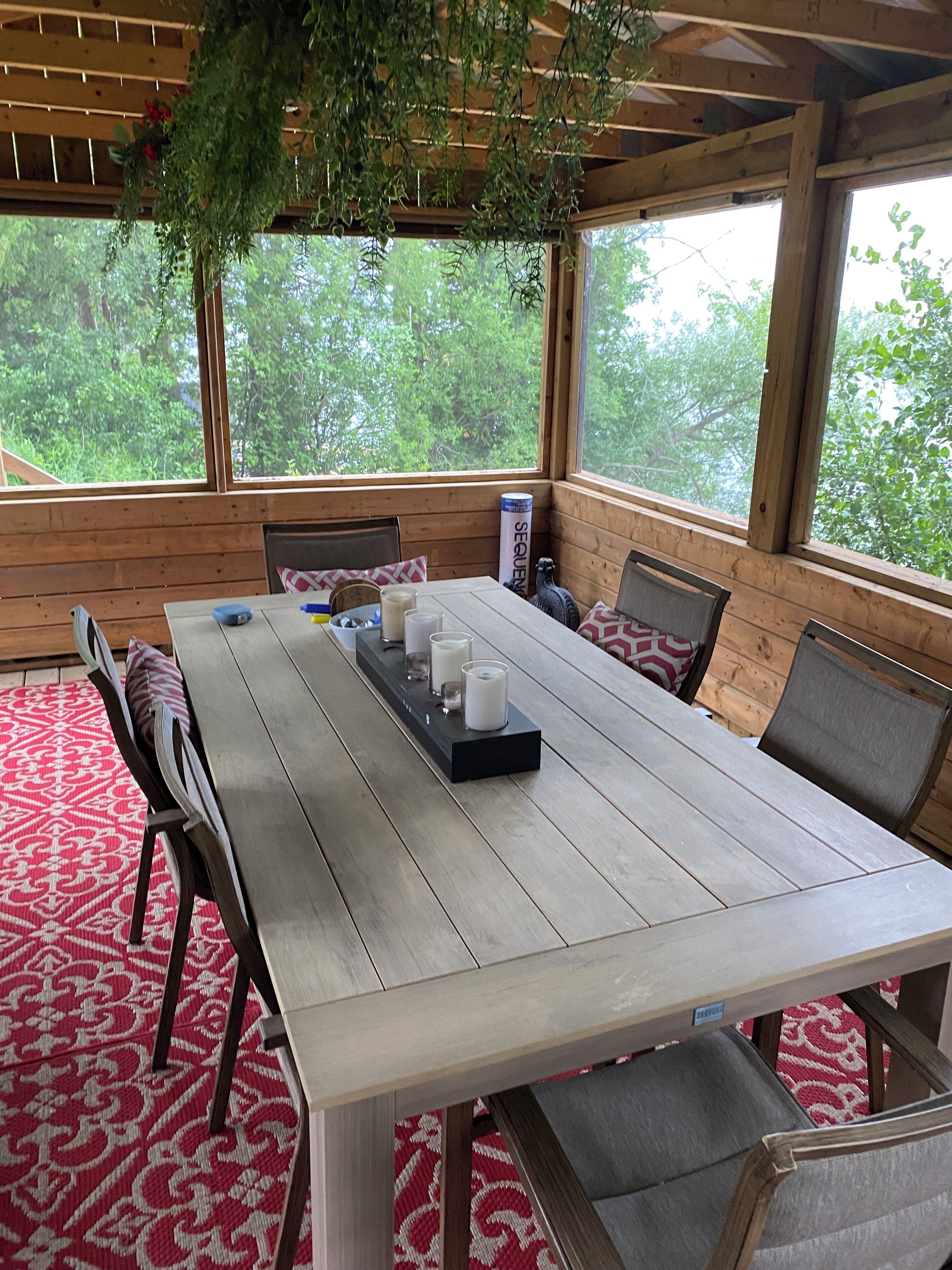 A wooden deck with a table and chairs, a hanging plant, and a view of trees.