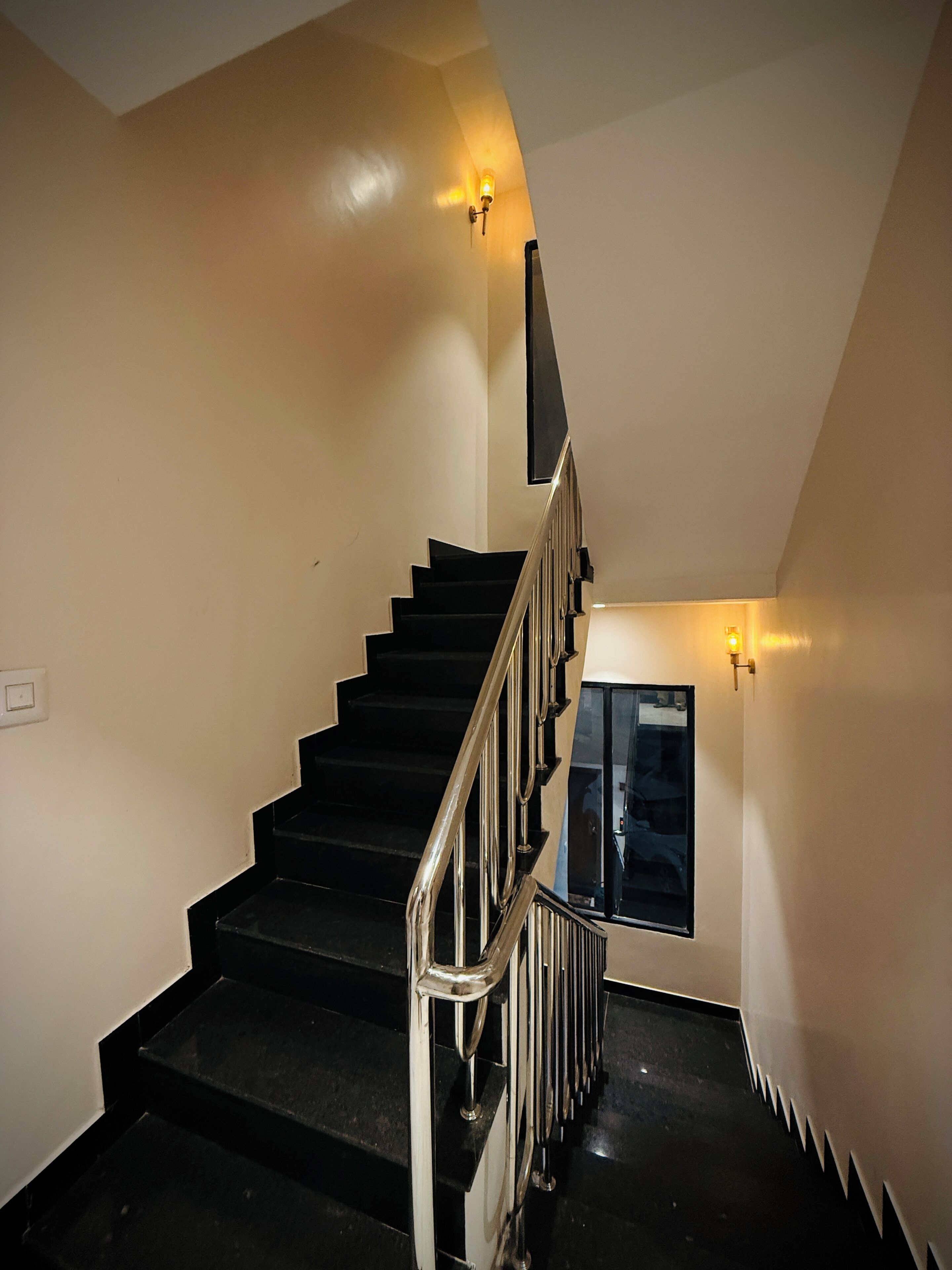 A staircase with black steps and a metal railing, flanked by light-colored walls and wall-mounted lights.
