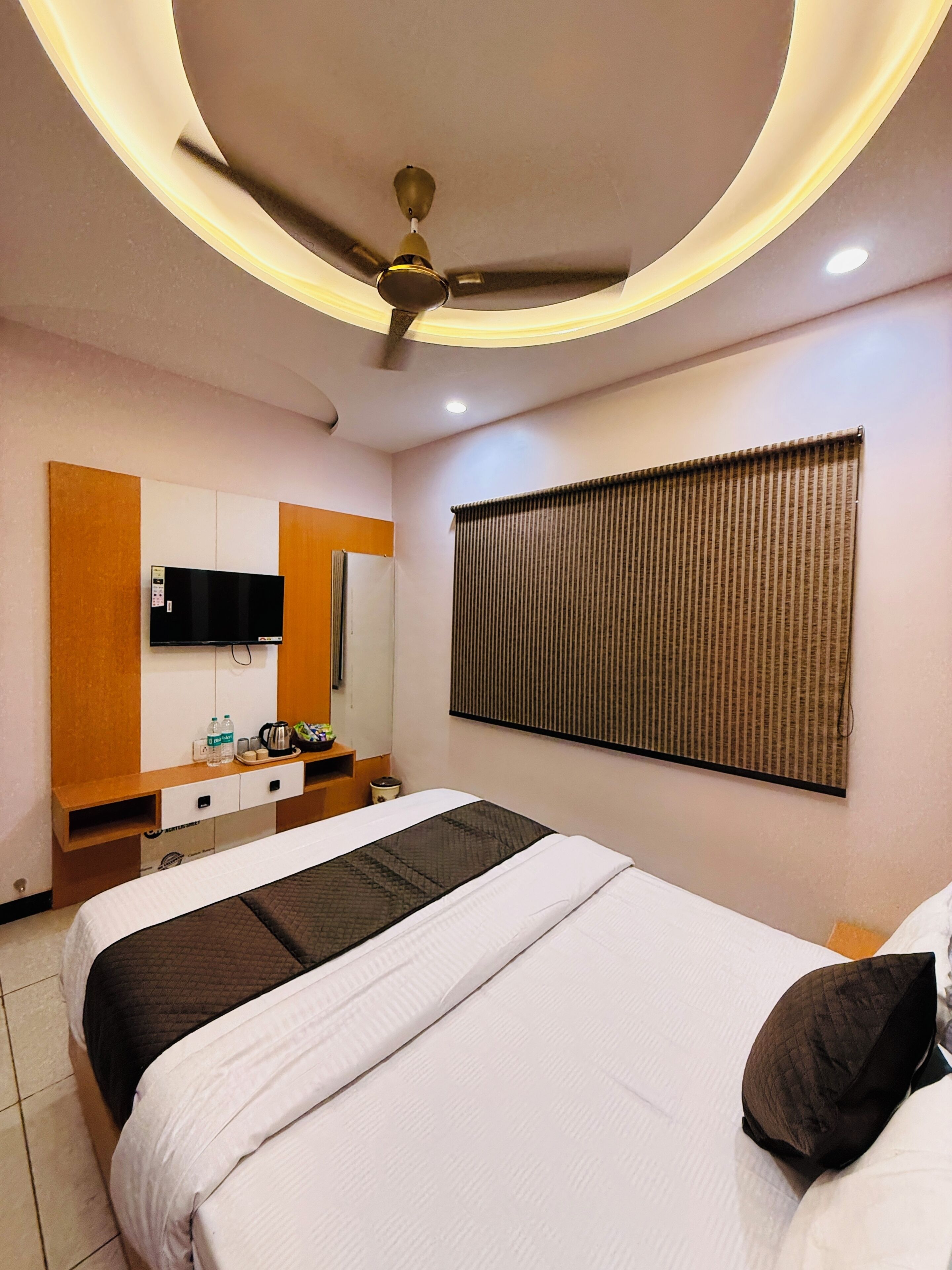 A hotel room with a bed, a flat-screen TV, a desk with a mirror, a window with blinds, and a ceiling fan.