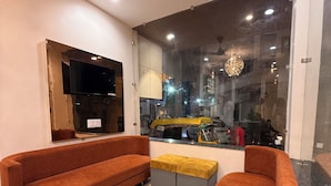 A modern living room with a sofa, ottoman, TV, and a large window with a view of the street. - Hotel Hira Palace (Ujjain)