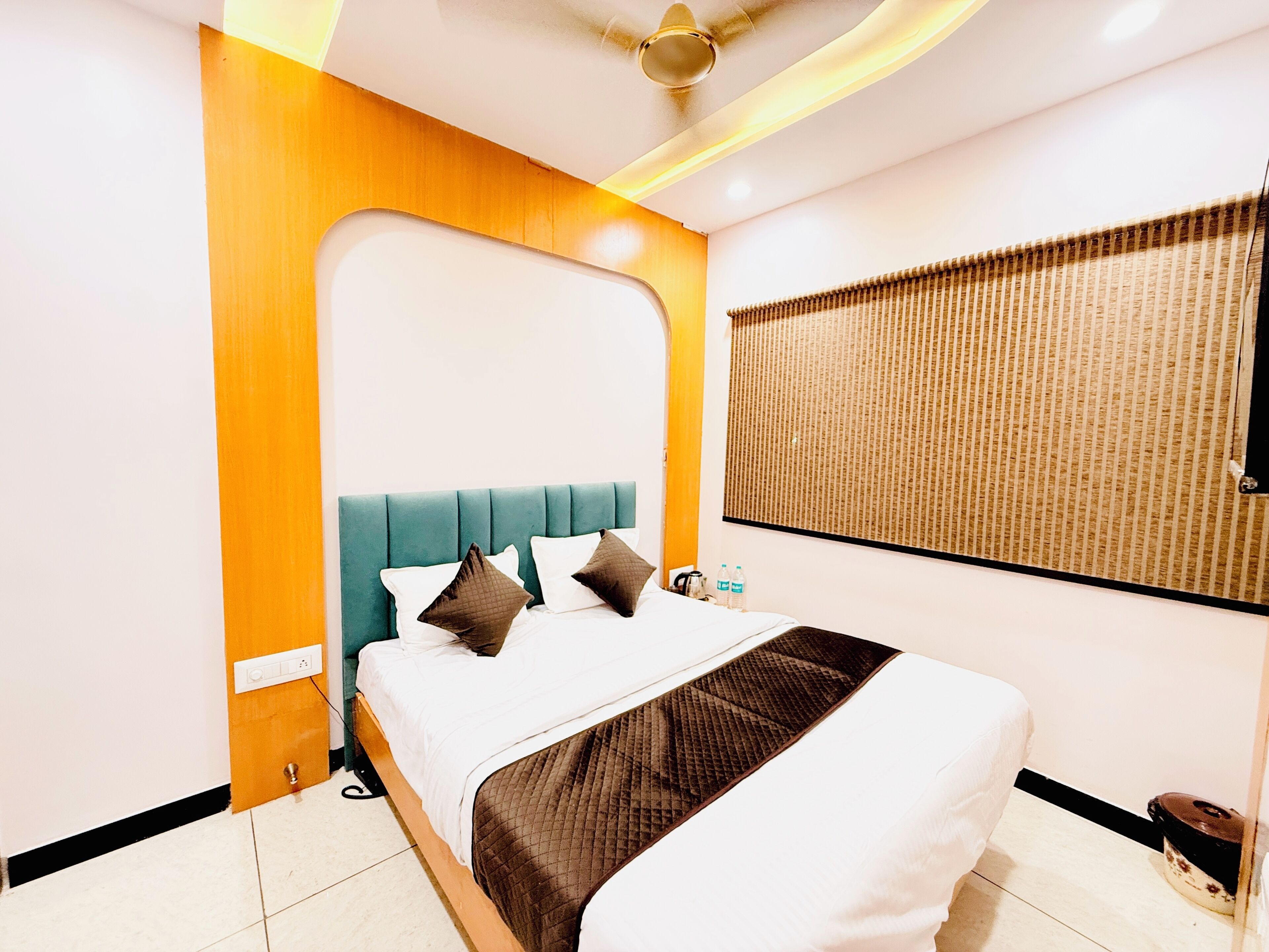 A modern hotel room with a teal headboard, white bedding, and a large window with a patterned blind.