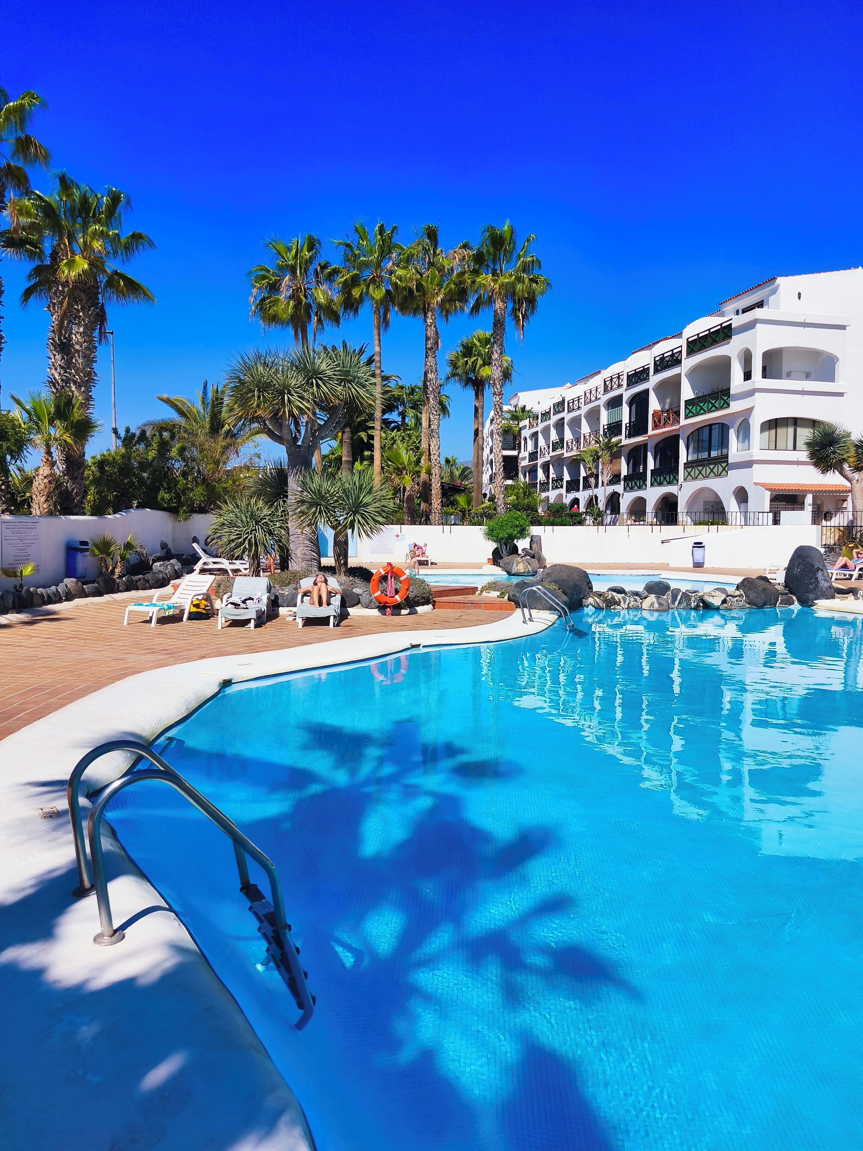 A swimming pool with lounge chairs, palm trees, and a multi-story building with balconies.