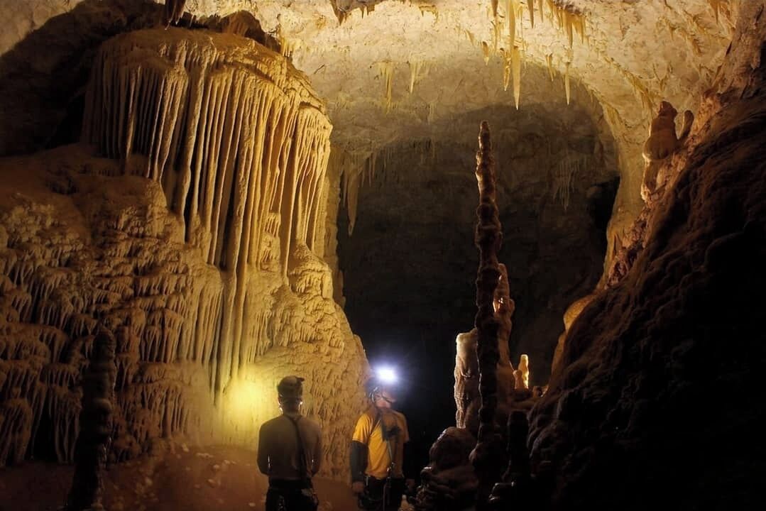 Una cueva con estalactitas y estalagmitas, una persona con linterna frontal y otra observando.