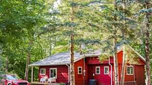 En rød hytte med veranda, en bil foran og skov i baggrunden.