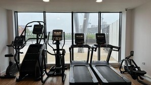 Un gimnasio con cintas de correr, elípticas y una bicicleta estática, con amplias ventanas y vistas a edificios.