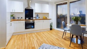 Una cocina moderna con armarios blancos, encimera de madera y horno. Cuenta con un comedor con mesa y sillas, y un amplio ventanal con vista a la ciudad.