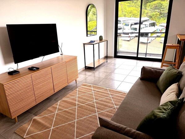 A living room with a TV on a wooden cabinet, a sofa, a small table, and a view of parked vehicles outside. - Sunset Moderne Apartment - Balconies - 5 min. from Marina du Marin (Le Marin)