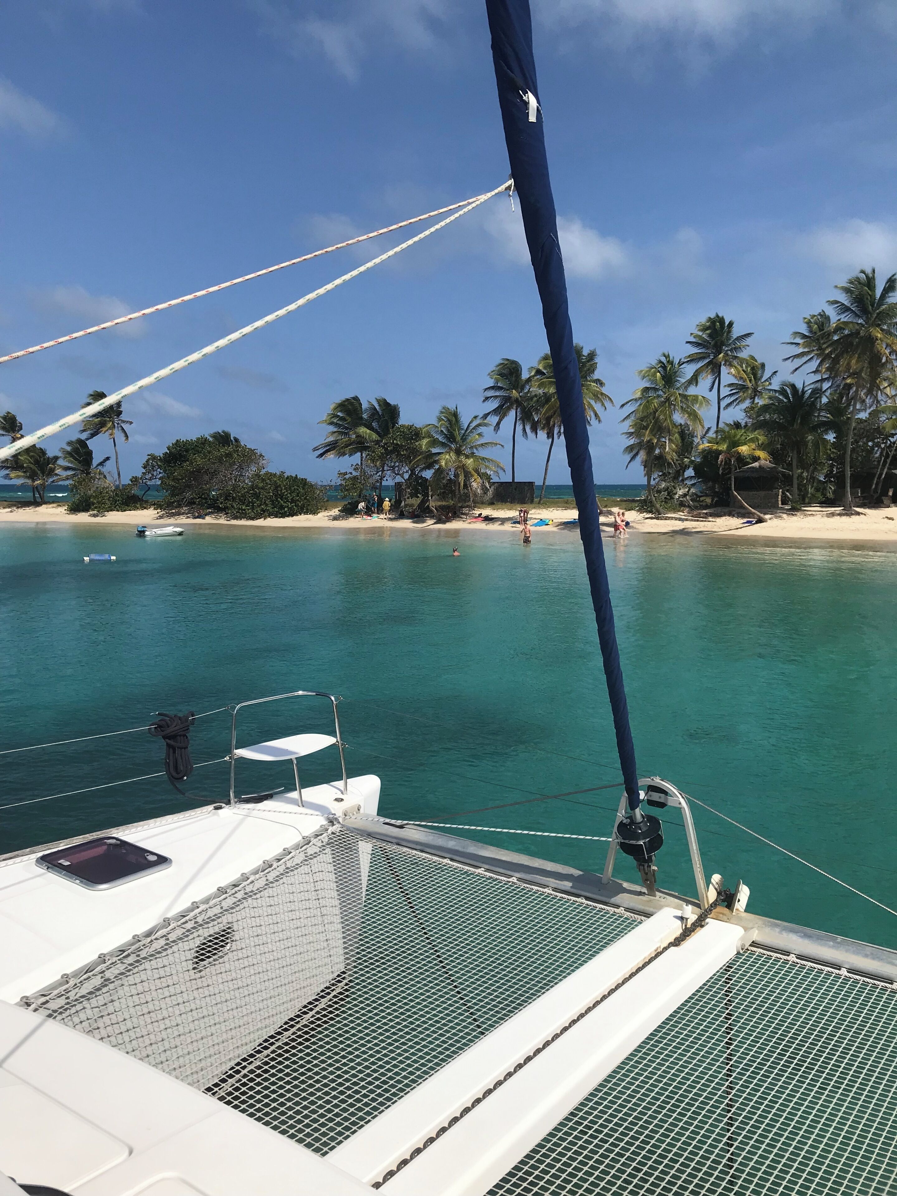 A boat with a netted deck and ropes, a clear turquoise sea, and a beach with palm trees and people in the background.