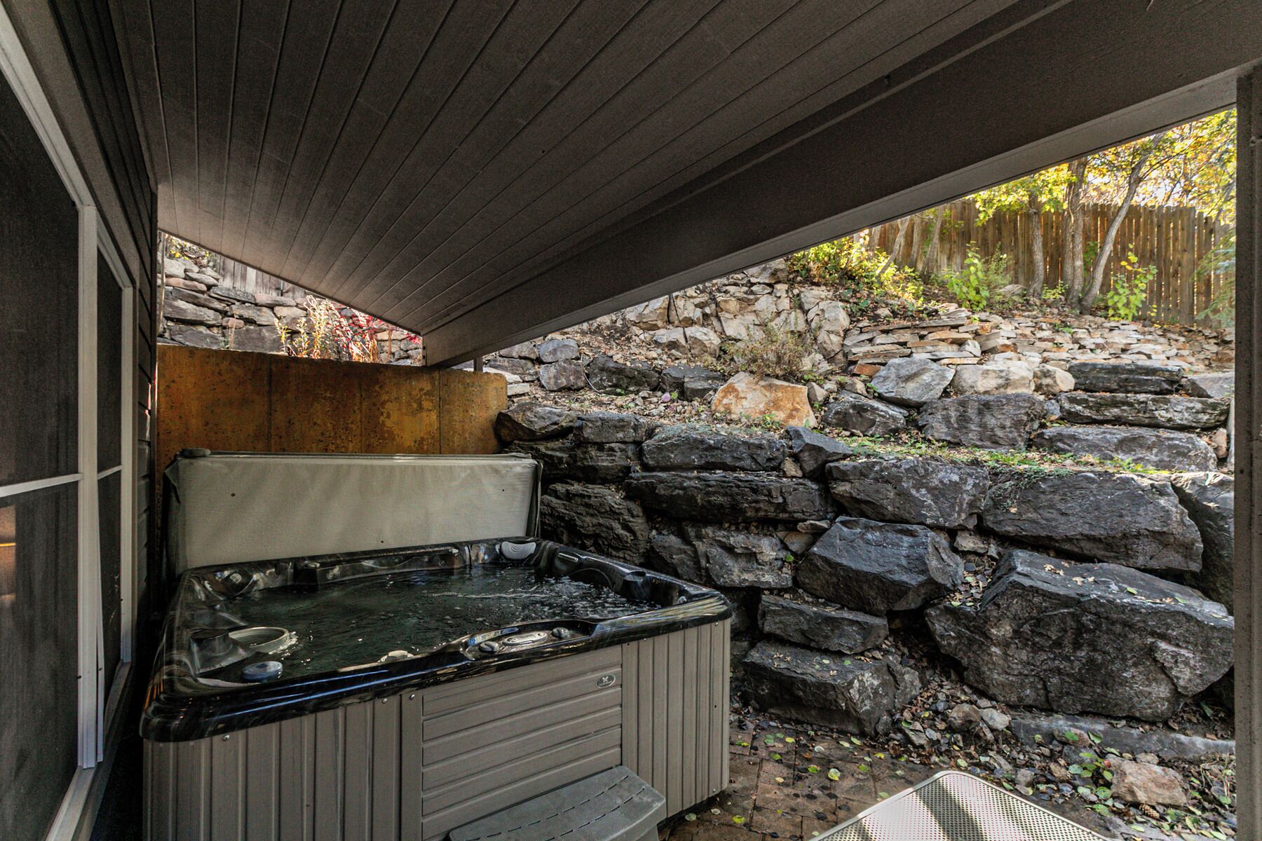 Un bain à remous sous une terrasse couverte, avec un mur en pierre et une clôture en bois en arrière-plan.