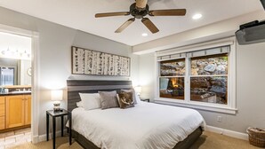 Une chambre avec un lit, un ventilateur de plafond, une fenêtre donnant sur des rochers et un coin cuisine visible à travers une porte ouverte.