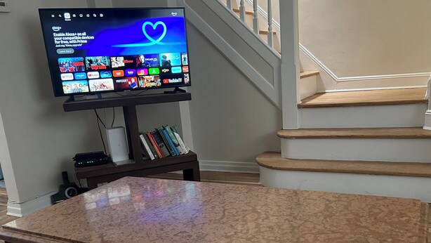 Ein Flachbildfernseher auf einem Standfuß, der einen Homescreen mit verschiedenen Streaming-Apps zeigt, ein Bücherregal mit Büchern und eine Treppe im Hintergrund.