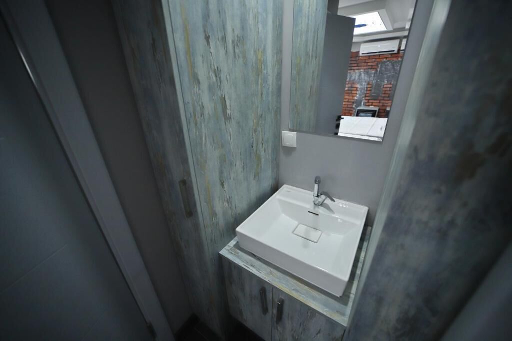 Un baño estrecho con un lavamanos blanco, un espejo y vista a una pared de ladrillo a través del ventanal.