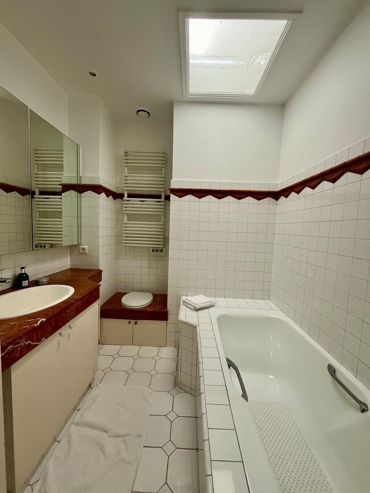 Un baño con lavamanos, inodoro y bañera, con azulejos blancos y un tragaluz.