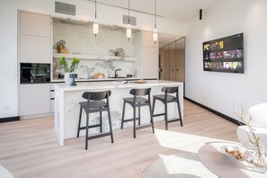A modern kitchen with a marble island, three bar stools, pendant lighting, and a wall-mounted TV displaying various shows.